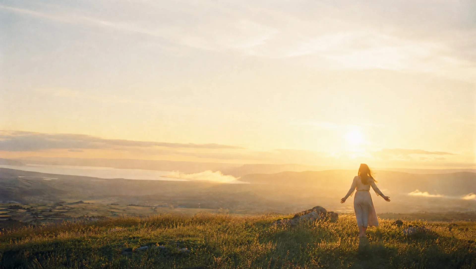 Eine Frau steht mit offenen Armen auf einer Hügelkuppe im Sonnenaufgang, vor ihr eine weite Landschaft mit Nebel im Tal